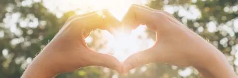Zwei Hände bilden mit den Finger ein Herz. Im Hintergrund strahlt die Sonne in Richtung Betrachter