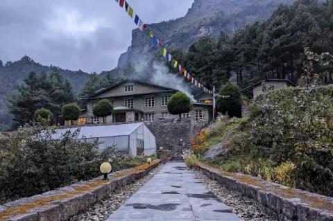 Ein Weg auf Steinplatten führt zu einem Haus. Im Hintergrund ist Wald und Nebel zu sehen. Über dem Haus weht eine buddhistische Gebetsfahne.
