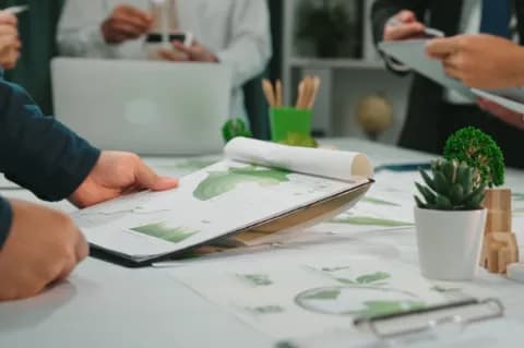 Szene einer Geschäftsbesprechung mit Fokus auf Nachhaltigkeit. Auf dem Tisch liegen Dokumente mit verschiedenen Diagrammen. Auf dem Tisch steht eine kleine Pflanze sowie weitere Büromaterialien.