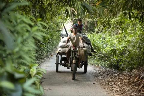 Ein Motorrad mit Anhänger fährt eine schmale Straße entlang. Auf dem Anhänger liegen weiße Säcke. Ein Mann fährt das Motorrad, der andere sitzt hinten auf den Säcken.