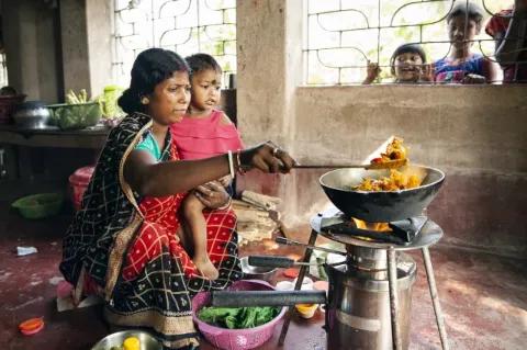 Eine Frau in traditionellen, indischem Gewand sitzt auf dem Boden und hebt mit einer Schöpfkelle etwas Essen aus einem Topf der über einem Holzofen steht. Auf ihrem Schoß sitzt ein Kind.