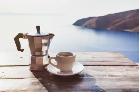 Ein Espressokocher und eine Espressotasse mit Kaffee stehen auf einem Holztisch vor einem See.