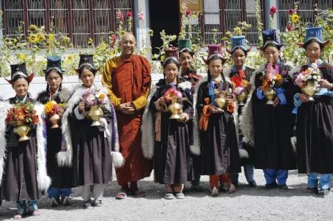 Ein Mönch in gelb-orangener Kutte steht in einer Gruppe von Nonnenschülerinnen vor einem Gebäude. Die Mädchen haben Vasen mit Blumen in den Händen.