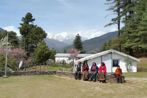 5 Menschen sitzen auf einer niedrigen Steinmauer vor zwei weiß gestrichenen Häusern und warten auf den Arztbesuch. Im Hintergrund sind Wald und Berge zu sehen.