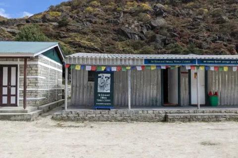 Ein kleines Gebäude aus Wellblech steht vor einem grünen Hügel. Am Dach hängen bunte, buddhistische Fahnen. Auf dem Schild vor dem Eingang steht "Sir Edmund Hillary Visitor Centre".