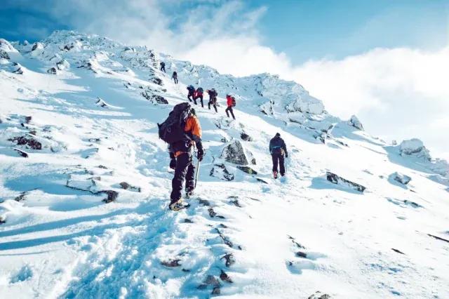 8 Personen laufen hintereinander einen verschneiten Berghang hoch bei blauem Himmel und Wolken