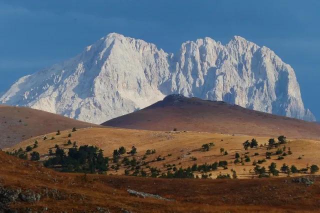 Ein großer, grau-weißer Berg am Horizont. Davor sind kleinere orange-braune Berge mit Bäumen.