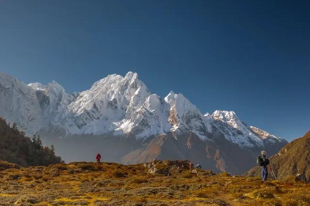 Zwei Wanderer blicken von einem orange-grün bewachsenen Berg auf einen schneebedeckten Berg.