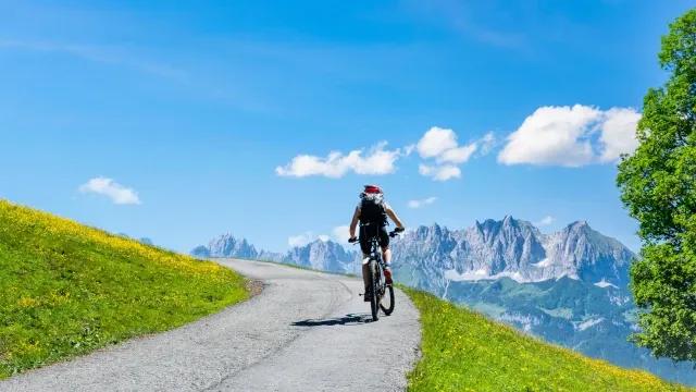 Eine Person auf dem Mountainbike fährt eine Teerstraße den Berg hoch. Neben ihm sind grüne Wiesen und über ihm ein strahlend blauer Himmel. Im Hintergrund sieht man Berge.
