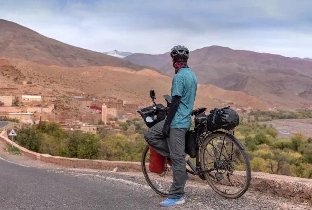 Ein Fahrradfahrer steht am Straßenrand mit seinem Fahrrad und blickt auf ein Dorf, braune Bergen und grüne Wälder.