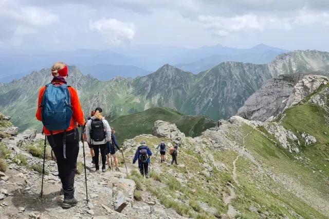 Sechs Personen mit Wanderstöcken laufen hintereinander über einen Grat bei bewölktem Himmel