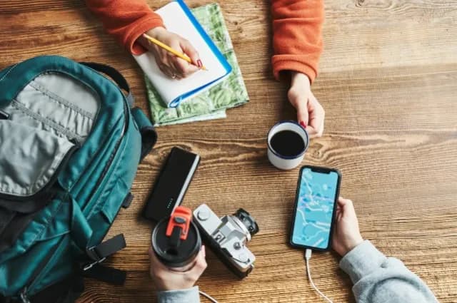 ein Tisch von oben mit Blick auf die Hände zweier Menschen. Auf dem Tisch liegen ein Handy, ein Rucksack, eine Kaffeetasse, ein Thermobecher, eine Karte und eine Kamera
