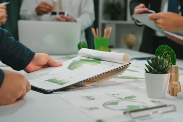 Szene einer Geschäftsbesprechung mit Fokus auf Nachhaltigkeit. Auf dem Tisch liegen Dokumente mit verschiedenen Diagrammen. Auf dem Tisch steht eine kleine Pflanze sowie weitere Büromaterialien.