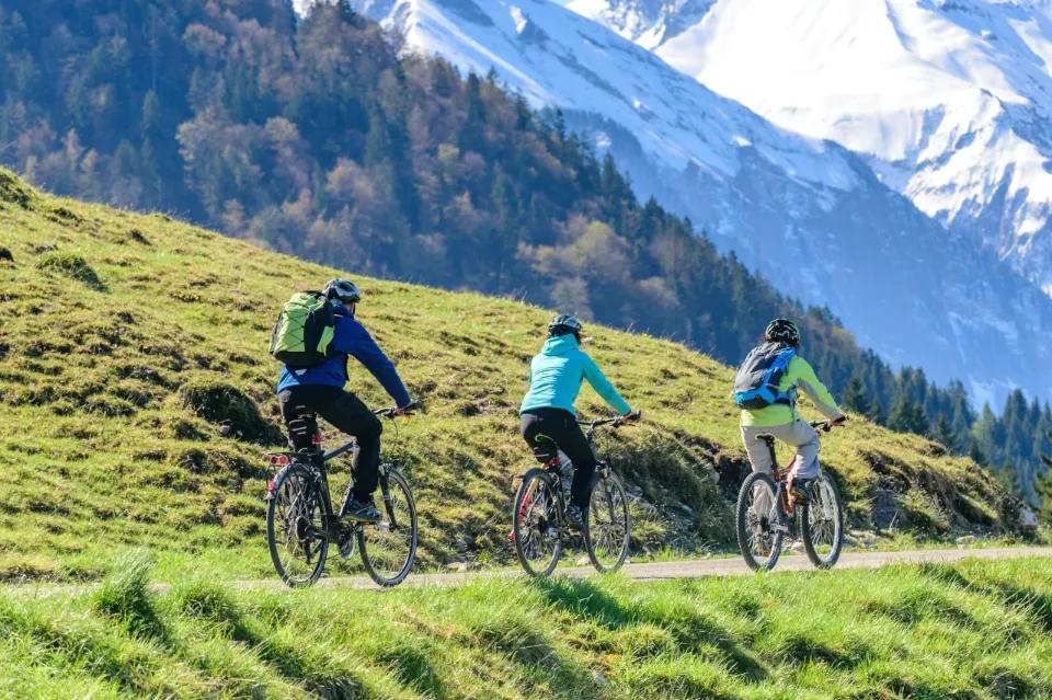 Drei Radfahrer fahren hintereinander auf einer Straße neben einer Wiese und vor hohen Bergen