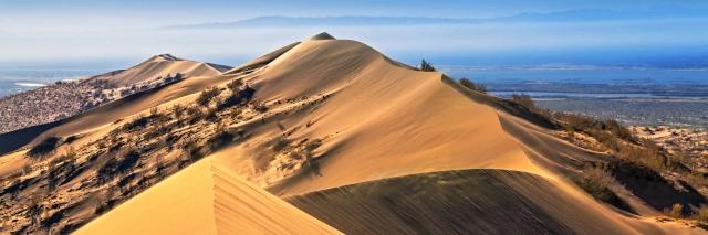 Hohe Sanddüne mit einzelnen Gewächsen führt Richtung blauem Horizont mit Wolken
