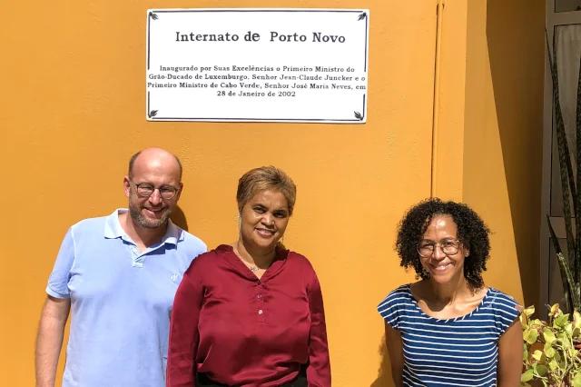 Drei Menschen stehen vor einem orangenen Gebäude und lachen in die Kamera.