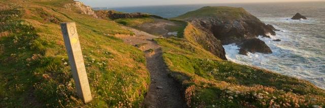 Wegweiser an einem Küstenpfad mit grüner Wiese und tosender Brandung bei Sonnenuntergang