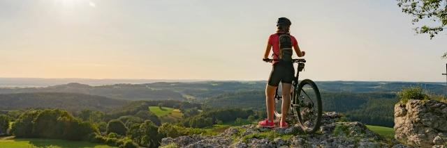 Eine junge Frau steht mit ihrem Mountainbike auf einem Aussichtspunkt und blickt über eine Wälder und Wiesen. Es ist ein trüber Himmel aber die Sonne scheint.