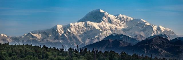 Blick auf einen schneebedeckten, felsigen Gebirgszug. Im Vordergrund sind die Gipfel eines Nadelwaldes zu sehen.