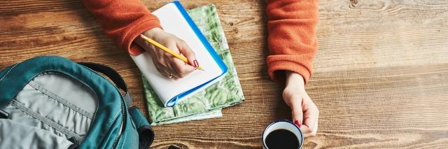 Auf einem Tisch liegt eine Landkarte und ein Notizbuch. Eine Frau hält einen Stift in der einen Hand und in der anderen eine Tasse Kaffee.