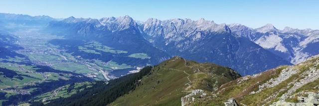 Panorama einer Berglandschaft mit grüner Wiese, bewaldeten Berghängen und grauen Bergspitzen