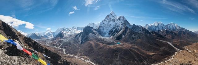 Panoramablick auf eine hochalpine Gebirgskette mit schneebedeckten Gipfeln. Vor der Gebirgskette ist ein tieferliegender Fluss zu sehen, der sich durch ein Tal schlägelt. Im Vordergrund weht eine buddhistische Gebetsflagge.