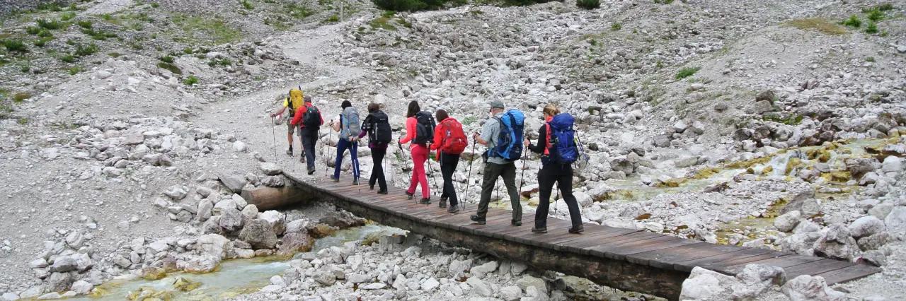 Eine Gruppe von acht Wanderern geht über eine kleine Holzbrücke. Der Untergrund ist felsig.