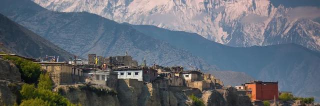 Blick auf ein kleines Dorf an felsigen Abgründen. Im Hintergrund ist das schneeweiße Nilgiri Himal zu sehen.