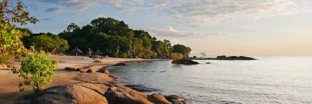 See mit Sandstrand, einzelnen Felsen, grünen Bäumen in einer Sonnenaufgangsstimmung