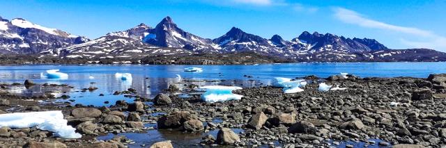 Fjord mit blauem Wasser, grauen Felsen und Eisböcken und Bergkette mit Schnee im Hintergrund