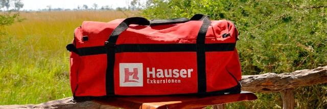 Eine rote Reisetasche mit Hauser Logo steht auf einem Holztisch.