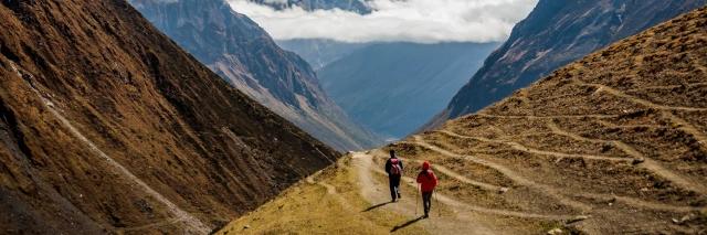 Zwei Wanderer auf Wanderweg, der durch ein Tal führt, mit schneebedeckten Bergen im Hintergrund