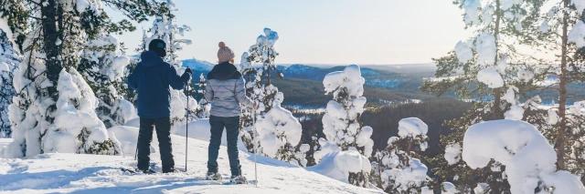 Zwei Wanderer mit Schneeschuhen und Wanderstöcken blicken über Schneelandschaft und Wald