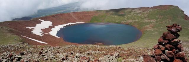 Ein See in einem großen Krater. Der Boden des Kraters ist grün, rot und teilweise schneebedeckt.