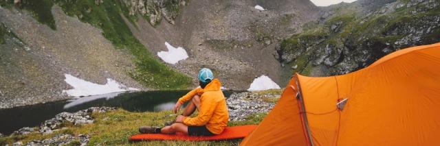 Ein Mann sitzt auf einer Isomatte vor seinem Zelt und schaut auf eine felsige Landschaft.