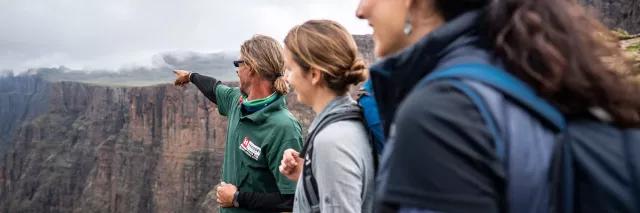 Reiseleiter zeigt mit dem Finger in die Ferne der Drakensberge in Südafrika und erklärt dabei zwei Personen etwas
