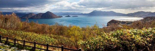 Küste mit braunen Sträuchern, einem blauen See und dunklen Bergen bei stark bewölktem Himmel