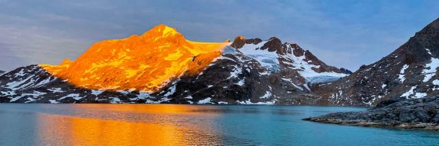 Am Rande des Wassers sind schneebedeckte Berge. Der Berg wird von der Sonne orange angestrahlt.