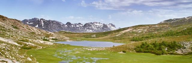 Landschaft mit grüner Wiese, grünem Berghang, See und Bergkette mit wenig Schnee im Hintergrund