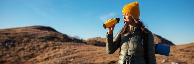 Eine junge Frau steht in der kargen Landschaft und trinkt Wasser aus einer Flasche.