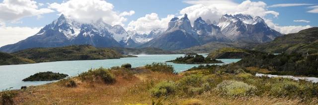 Ein blauer See. Im Hintergrund sind hohe schneebedeckte Berge, in denen die Wolken hängen.