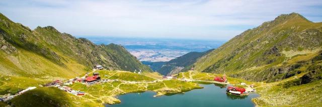 Landschaft mit blauem Bergsee, einzelnen Häusern und grünen Berghängen unter bewölktem Himmel