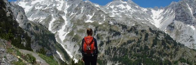 Eine Wanderin mit rotem Rucksack steht vor einem gräulichen, teils schneebdeckten Bergmassiv.