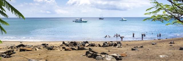 Ein Sandstrand und das blaue Meer mit Segelbooten. Im Wasser stehen mehrere Menschen.