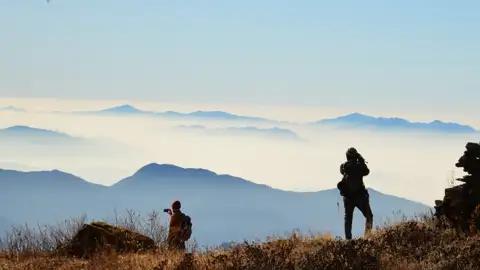 Zwei Wanderer mit Rucksäcken auf einem Berggipfel, die die Aussicht auf die nebligen Berge fotografieren.