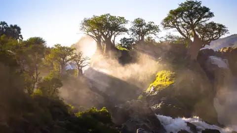 Sonnenaufgang über einem Wasserfall mit Baobab Bäumen im Vordergrund.