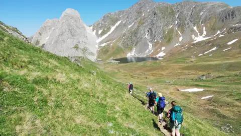 Wanderer auf einem Bergpfad inmitten einer Berglandschaft mit See und Gipfeln.