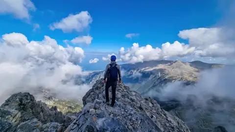 Wanderer auf einem Berggipfel mit Blick auf eine Berglandschaft und Wolken.
