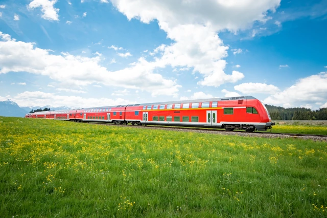 Ein roter Zug fährt durch eine grüne Landschaft. Am Himmel sind weiße Wolken. 