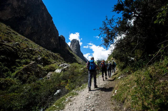 Eine Wandergruppe mit Stücken geht auf einem Schotterpfad entlang eines grünen Tals. 